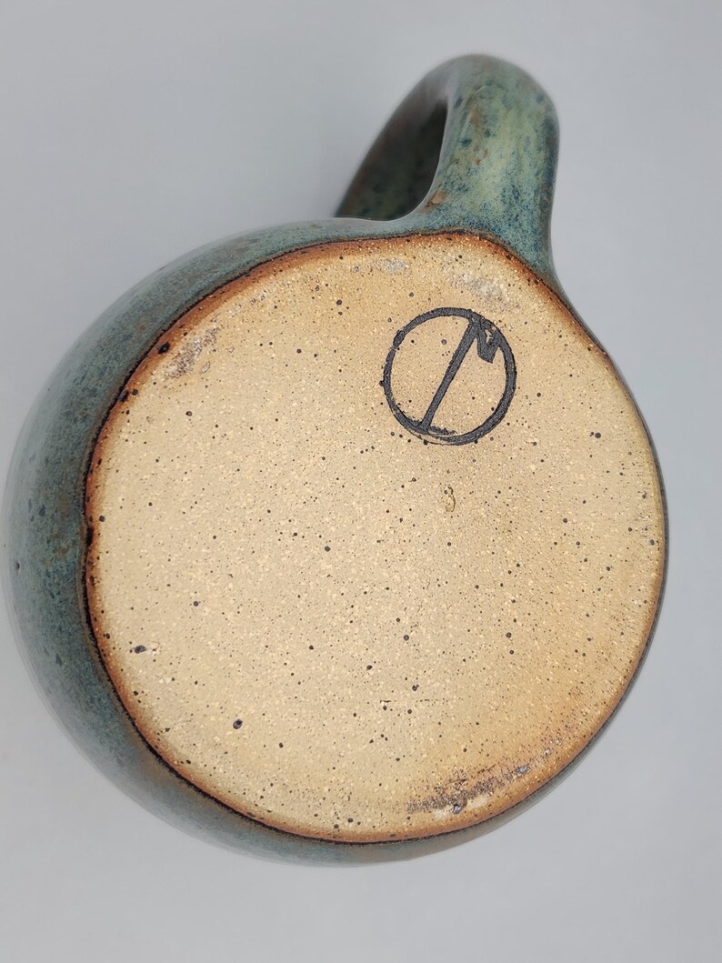 May include: A handmade ceramic mug with a rounded body and a handle. The mug has a speckled, light brown base and a teal-coloured exterior with a darker rim. A black circular stamp is visible on the base.