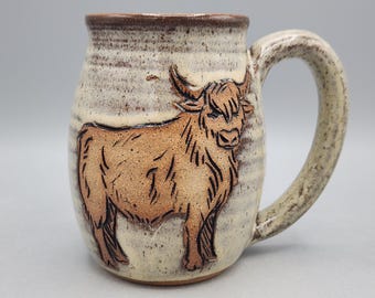 May include: A ceramic mug with a speckled cream and brown glaze featuring a raised, carved relief of a Highland cow in a tan color. The mug has a sturdy handle and a rounded body, showcasing a textured finish throughout the exterior surface.
