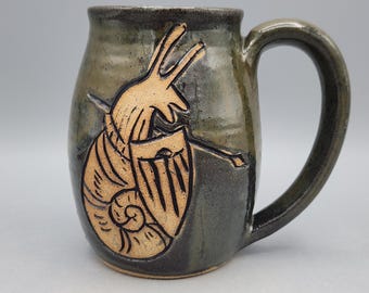 May include: A dark olive green ceramic mug featuring a carved, light tan graphic of a snail holding a shield and a spear. The mug has a smooth, glossy finish with a curved handle and a rounded body, showcasing a detailed relief design.