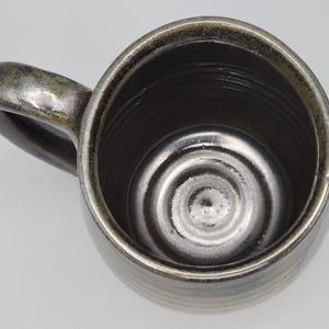 Puede incluir: Vista superior de una taza de cer&aacute;mica hecha a mano con un esmalte met&aacute;lico oscuro. La taza tiene un cuerpo redondeado, un asa robusta y una superficie texturizada. El interior presenta c&iacute;rculos conc&eacute;ntricos en la base.