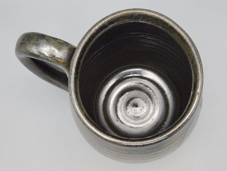 Puede incluir: Vista superior de una taza de cer&aacute;mica hecha a mano con un esmalte met&aacute;lico oscuro. La taza tiene un cuerpo redondeado, un asa robusta y una superficie texturizada. El interior presenta c&iacute;rculos conc&eacute;ntricos en la base.