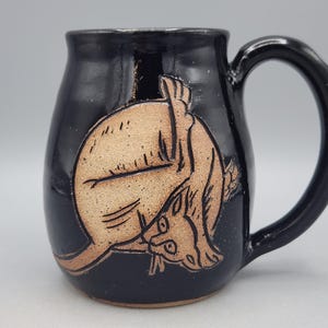 Może przedstawiać: Błyszczący czarny kubek ceramiczny z jasnobrązową, wygrawerowaną ilustracją kota zwiniętego w koło. Kot jest przedstawiony w pozycji podczas mycia się, z głową schowaną blisko tylnej łapy, co ukazuje precyzyjne wykonanie detali.
