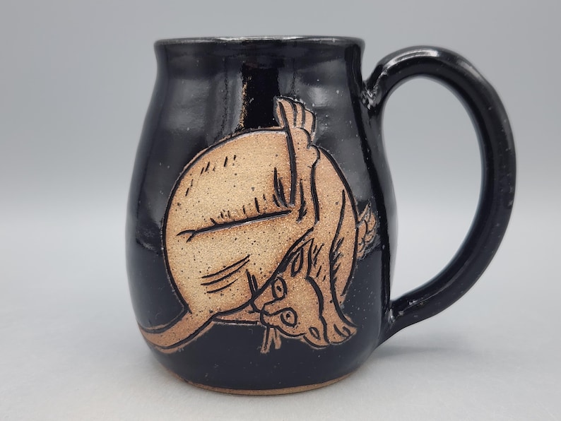 Może przedstawiać: Błyszczący czarny kubek ceramiczny z jasnobrązową, wygrawerowaną ilustracją kota zwiniętego w koło. Kot jest przedstawiony w pozycji podczas mycia się, z głową schowaną blisko tylnej łapy, co ukazuje precyzyjne wykonanie detali.