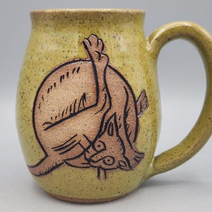 Może przedstawiać: Nakrapiany ż&oacute;łto-zielony ceramiczny kubek z ciemnobrązową wypukłą ilustracją kota myjącego się z uniesioną nogą. Kubek ma zaokrąglony korpus, szeroki brzeg oraz zakrzywione ucho znajdujące się po prawej stronie naczynia.