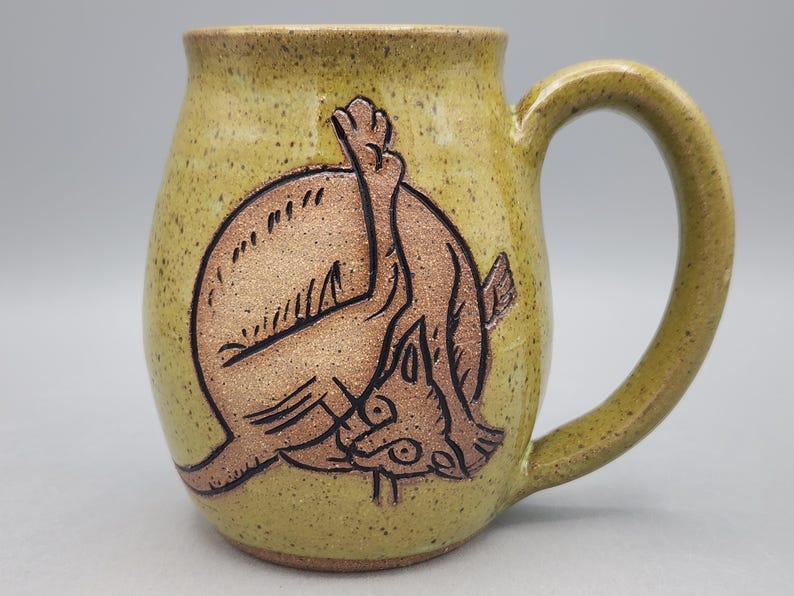 Może przedstawiać: Nakrapiany ż&oacute;łto-zielony ceramiczny kubek z ciemnobrązową wypukłą ilustracją kota myjącego się z uniesioną nogą. Kubek ma zaokrąglony korpus, szeroki brzeg oraz zakrzywione ucho znajdujące się po prawej stronie naczynia.