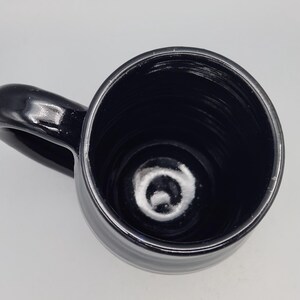 Puede incluir: Una taza de cer&aacute;mica negra brillante con un asa curva. El interior de la taza tambi&eacute;n es negro y la taza est&aacute; vac&iacute;a. La taza se ve desde arriba, mostrando el interior y el asa.