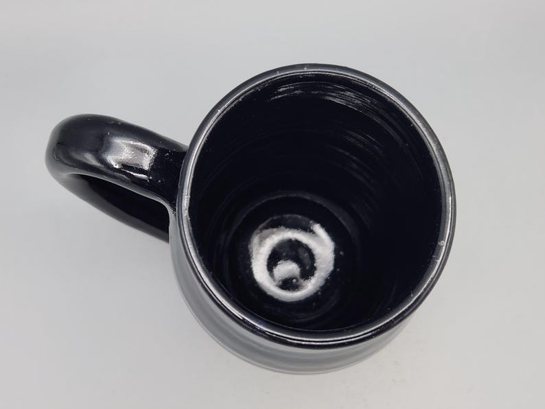 Puede incluir: Una taza de cer&aacute;mica negra brillante con un asa curva. El interior de la taza tambi&eacute;n es negro y la taza est&aacute; vac&iacute;a. La taza se ve desde arriba, mostrando el interior y el asa.