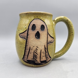 May include: A ceramic mug with a speckled olive green glaze featuring a carved, light brown ghost figure on the front. The ghost has two dark oval eyes and an open, oval-shaped mouth. The mug has a rounded body and a curved handle on the right side.