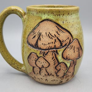 Puede incluir: Una taza de cer&aacute;mica hecha a mano con un dise&ntilde;o caprichoso de hongos. La taza presenta un esmalte moteado verde claro y amarillo, con ilustraciones de hongos marrones. El asa es de color verde claro a juego. Una pieza &uacute;nica para caf&eacute; o t&eacute;.