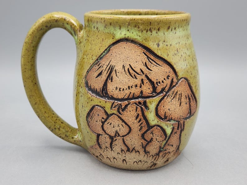 Puede incluir: Una taza de cer&aacute;mica hecha a mano con un dise&ntilde;o caprichoso de hongos. La taza presenta un esmalte moteado verde claro y amarillo, con ilustraciones de hongos marrones. El asa es de color verde claro a juego. Una pieza &uacute;nica para caf&eacute; o t&eacute;.