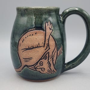 Może przedstawiać: Ciemnozielony ceramiczny kubek z wygrawerowaną, nieszkliwioną ilustracją kota w kolorze opalonej gliny, kt&oacute;ry myje się z uniesioną nogą. Kubek ma błyszczące wykończenie, zaokrąglony korpus oraz zakrzywione ucho po prawej stronie naczynia.