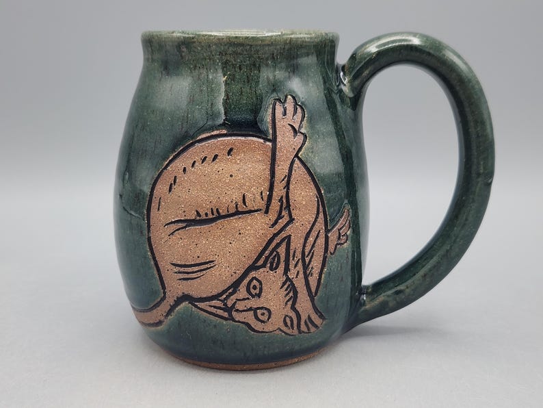 Może przedstawiać: Ciemnozielony ceramiczny kubek z wygrawerowaną, nieszkliwioną ilustracją kota w kolorze opalonej gliny, kt&oacute;ry myje się z uniesioną nogą. Kubek ma błyszczące wykończenie, zaokrąglony korpus oraz zakrzywione ucho po prawej stronie naczynia.