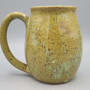 Może przedstawiać: Ceramiczny kubek o zaokrąglonym korpusie i zakrzywionym uchwycie, pokryty nakrapianą musztardowo-ż&oacute;łtą i jasnozieloną glazurą. Powierzchnia ma teksturę z ciemnobrązowymi plamkami rozłożonymi na zewnętrznej stronie naczynia.