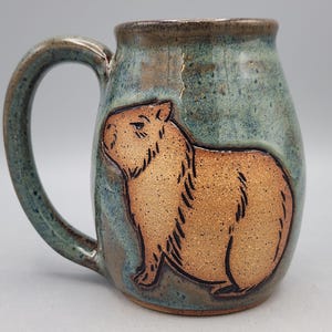 May include: A ceramic mug with a capybara design. The mug has a brown capybara illustration on a teal and brown background. The handle and rim are a darker teal colour. A unique, handcrafted piece.