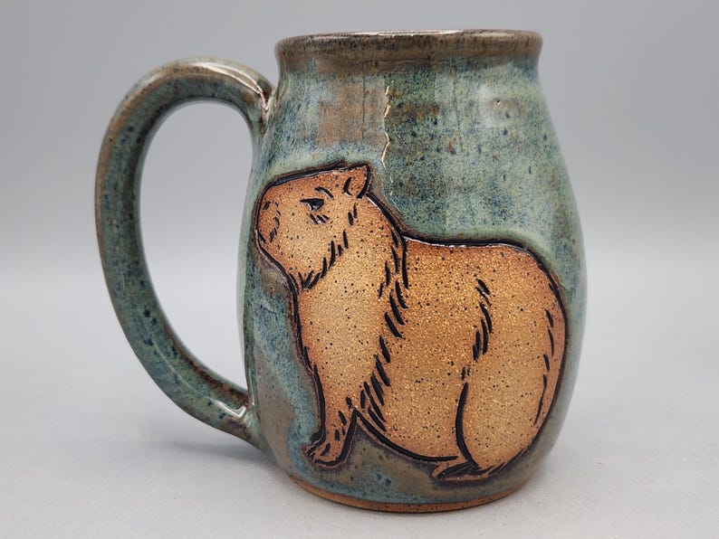 May include: A ceramic mug with a capybara design. The mug has a brown capybara illustration on a teal and brown background. The handle and rim are a darker teal colour. A unique, handcrafted piece.