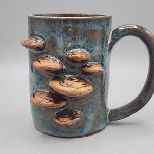 Może przedstawiać: Ceramiczny kubek z niebiesko-brązowym nakrapianym szkliwem, ozdobiony kilkoma wypukłymi, teksturowanymi brązowymi naroślami przypominającymi grzyby na zewnętrznej powierzchni. Kubek posiada solidne, zakrzywione ucho i cylindryczny korpus.
