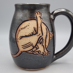 May include: A dark charcoal-colored ceramic mug with a glossy finish and a large, curved handle. The front features a light tan, etched illustration of a cat curled into a ball, grooming itself with one leg extended upward toward its back.