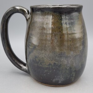 Puede incluir: Una taza de cer&aacute;mica hecha a mano con un esmalte met&aacute;lico oscuro. La taza tiene un cuerpo redondeado y un asa grande y curvada. Los colores van del gris oscuro en la base a un tono m&aacute;s claro, similar al bronce, hacia la parte superior. Para bebidas fr&iacute;as o calientes.