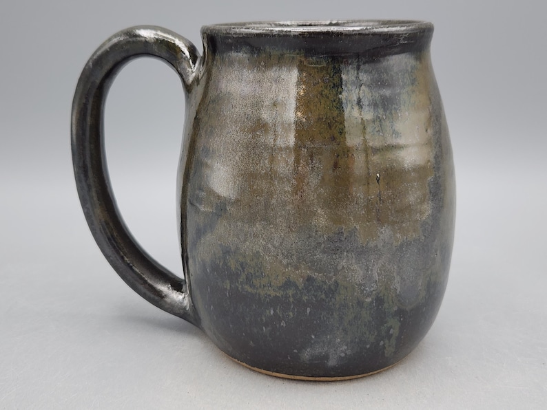 Puede incluir: Una taza de cer&aacute;mica hecha a mano con un esmalte met&aacute;lico oscuro. La taza tiene un cuerpo redondeado y un asa grande y curvada. Los colores van del gris oscuro en la base a un tono m&aacute;s claro, similar al bronce, hacia la parte superior. Para bebidas fr&iacute;as o calientes.