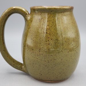 Puede incluir: Una taza de cer&aacute;mica hecha a mano con un cuerpo redondeado y un asa grande y curvada. La taza tiene un esmalte verde oliva moteado con detalles marrones. El interior y el borde son de un tono marr&oacute;n m&aacute;s oscuro. La taza mide unos 10 cm de alto.