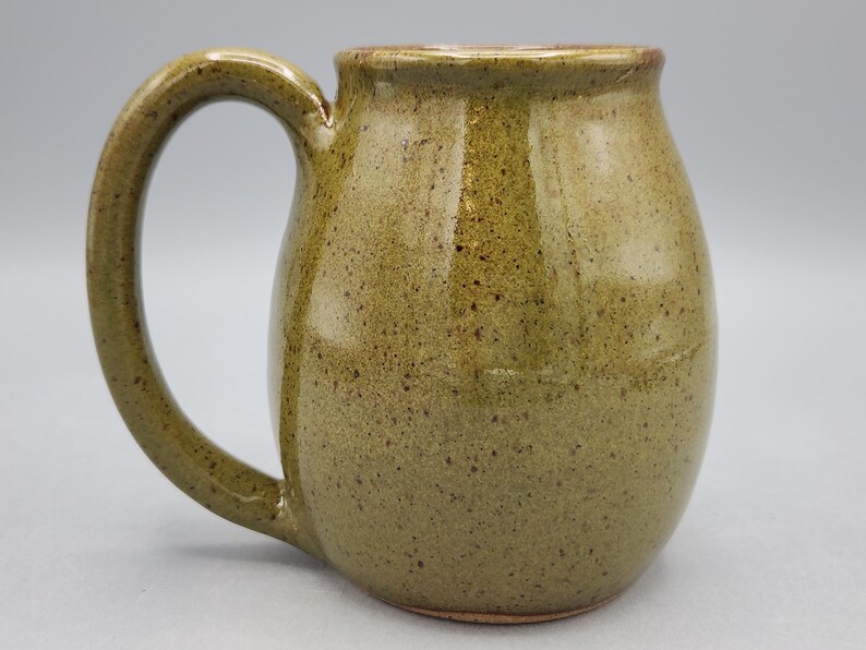 Puede incluir: Una taza de cer&aacute;mica hecha a mano con un cuerpo redondeado y un asa grande y curvada. La taza tiene un esmalte verde oliva moteado con detalles marrones. El interior y el borde son de un tono marr&oacute;n m&aacute;s oscuro. La taza mide unos 10 cm de alto.