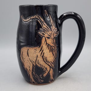 Puede incluir: Taza de cer&aacute;mica negra con un asa grande y curvada. La taza presenta una ilustraci&oacute;n detallada de una cabra de color marr&oacute;n claro. La cabra est&aacute; centrada en el cuerpo de la taza. La taza tiene un acabado brillante.