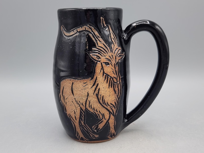 Puede incluir: Taza de cer&aacute;mica negra con un asa grande y curvada. La taza presenta una ilustraci&oacute;n detallada de una cabra de color marr&oacute;n claro. La cabra est&aacute; centrada en el cuerpo de la taza. La taza tiene un acabado brillante.