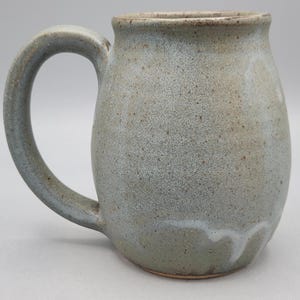 Może przedstawiać: Ceramiczny kubek o zaokrąglonym korpusie i dużym uchwycie. Powierzchnia pokryta jest nakrapianym jasnoszarym i niebieskim szkliwem o gładkim wykończeniu. U podstawy widoczny jest subtelny biały wz&oacute;r zaciek&oacute;w, a krawędź jest brązowa.