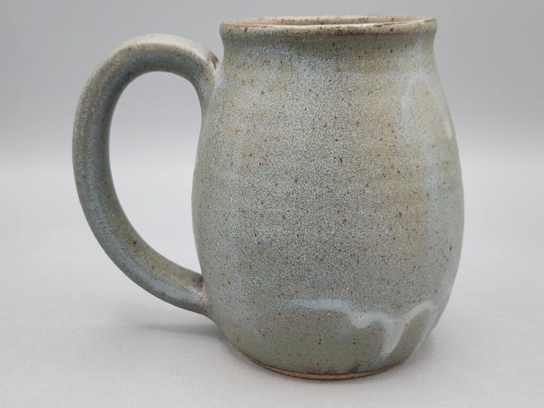 Może przedstawiać: Ceramiczny kubek o zaokrąglonym korpusie i dużym uchwycie. Powierzchnia pokryta jest nakrapianym jasnoszarym i niebieskim szkliwem o gładkim wykończeniu. U podstawy widoczny jest subtelny biały wz&oacute;r zaciek&oacute;w, a krawędź jest brązowa.