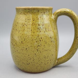 May include: A handmade ceramic mug with a speckled, olive-green glaze. The mug has a rounded body, a wide handle, and a light brown rim. The mug is designed for hot beverages.
