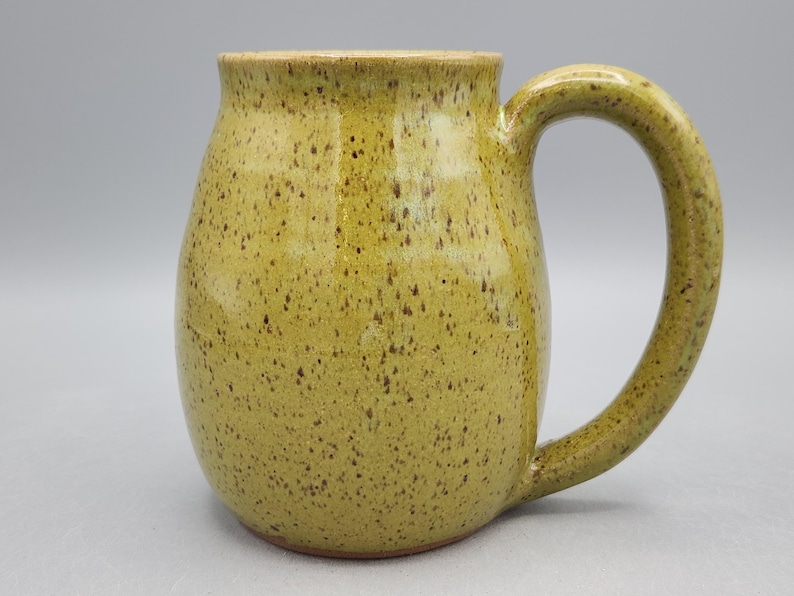 May include: A handmade ceramic mug with a speckled, olive-green glaze. The mug has a rounded body, a wide handle, and a light brown rim. The mug is designed for hot beverages.