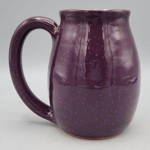 May include: A handcrafted ceramic mug in a deep purple colour with a speckled finish. The mug has a large, curved handle and a rounded body. The interior is a lighter shade of purple. Ideal for tea, coffee, or other drinks.