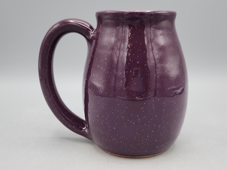 May include: A handcrafted ceramic mug in a deep purple colour with a speckled finish. The mug has a large, curved handle and a rounded body. The interior is a lighter shade of purple. Ideal for tea, coffee, or other drinks.
