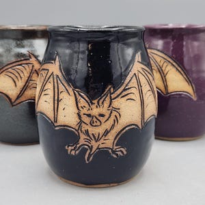 May include: Three ceramic mugs with handles, each featuring a carved bat design. The centre mug is glossy black, the left mug is metallic charcoal, and the right mug is deep purple. Each bat is rendered in a natural tan clay colour with detailed wings.