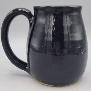 Może przedstawiać: Ręcznie wykonany ceramiczny kubek z błyszczącym, głębokim czarnym szkliwem i zaokrąglonym, pękatym korpusem. Kubek posiada duże, zakrzywione ucho po lewej stronie oraz gładką, odblaskową powierzchnię z subtelnymi zmianami w ciemnym wykończeniu.