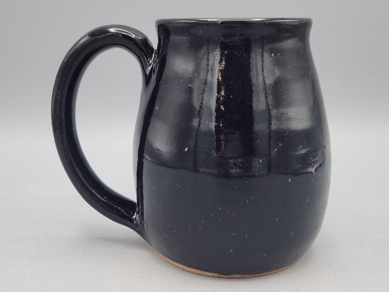 Może przedstawiać: Ręcznie wykonany ceramiczny kubek z błyszczącym, głębokim czarnym szkliwem i zaokrąglonym, pękatym korpusem. Kubek posiada duże, zakrzywione ucho po lewej stronie oraz gładką, odblaskową powierzchnię z subtelnymi zmianami w ciemnym wykończeniu.