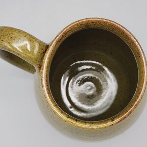 Puede incluir: Vista superior de una taza de cer&aacute;mica hecha a mano. La taza es de color verde oliva apagado con una forma redondeada y un asa grande y curvada. El borde y el asa tienen un acento marr&oacute;n moteado m&aacute;s oscuro. El interior de la taza es de un tono verde m&aacute;s claro.