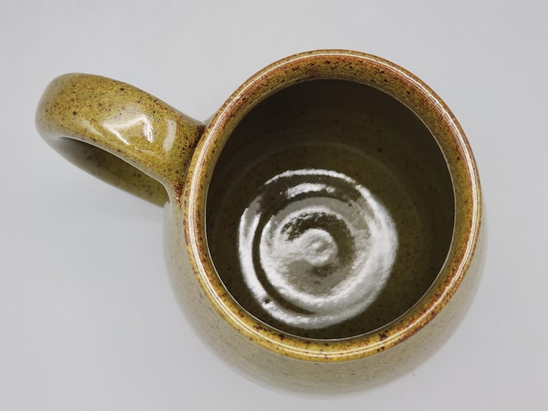 Puede incluir: Vista superior de una taza de cer&aacute;mica hecha a mano. La taza es de color verde oliva apagado con una forma redondeada y un asa grande y curvada. El borde y el asa tienen un acento marr&oacute;n moteado m&aacute;s oscuro. El interior de la taza es de un tono verde m&aacute;s claro.