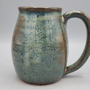 May include: A handmade ceramic mug with a rounded body and a large handle. The mug has a speckled blue-green glaze with brown accents. The rim and handle are a darker brown. Ideal for tea or coffee, this mug is a unique piece.
