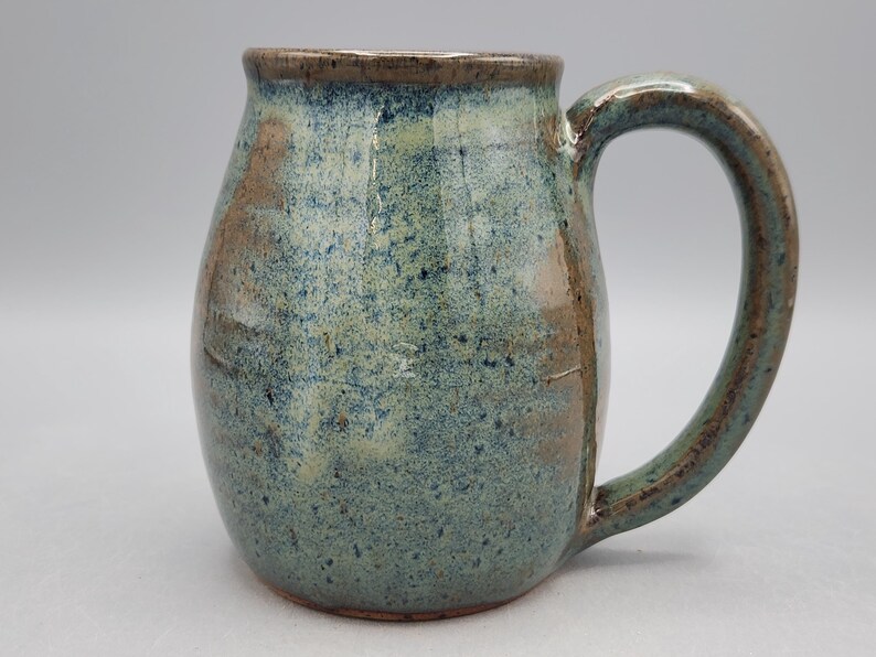 May include: A handmade ceramic mug with a rounded body and a large handle. The mug has a speckled blue-green glaze with brown accents. The rim and handle are a darker brown. Ideal for tea or coffee, this mug is a unique piece.