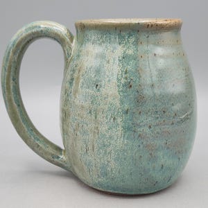 Może przedstawiać: Ceramiczny kubek o zaokrąglonym korpusie i dużym uchwycie. Powierzchnia pokryta jest nakrapianym szkliwem w odcieniach morskiej zieleni, turkusu i ziemistego brązu, z gładkim, błyszczącym wykończeniem i subtelnymi r&oacute;żnicami w kolorystyce.