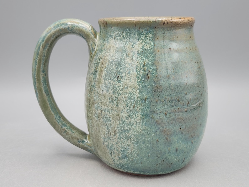 Może przedstawiać: Ceramiczny kubek o zaokrąglonym korpusie i dużym uchwycie. Powierzchnia pokryta jest nakrapianym szkliwem w odcieniach morskiej zieleni, turkusu i ziemistego brązu, z gładkim, błyszczącym wykończeniem i subtelnymi r&oacute;żnicami w kolorystyce.
