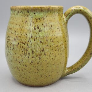Puede incluir: Una taza de cer&aacute;mica hecha a mano con un cuerpo redondeado y un asa grande y curvada. La taza tiene un aspecto moteado y terroso con tonos de verde oliva y marr&oacute;n. La taza est&aacute; dise&ntilde;ada para bebidas calientes.