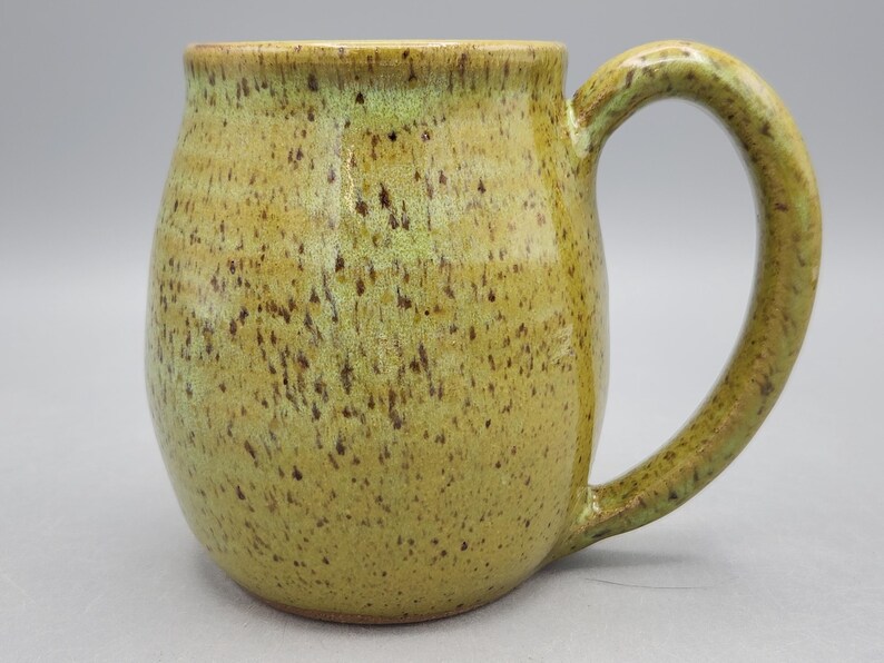Puede incluir: Una taza de cer&aacute;mica hecha a mano con un cuerpo redondeado y un asa grande y curvada. La taza tiene un aspecto moteado y terroso con tonos de verde oliva y marr&oacute;n. La taza est&aacute; dise&ntilde;ada para bebidas calientes.