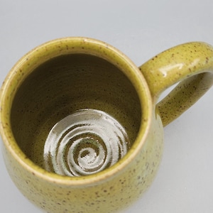 May include: A handmade ceramic mug with a speckled, yellowish-green glaze. The mug has a rounded body, a large handle, and a spiral design at the bottom. The interior is a darker shade of green.