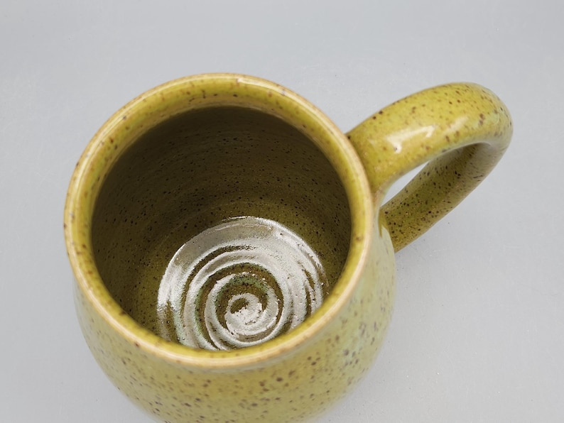 May include: A handmade ceramic mug with a speckled, yellowish-green glaze. The mug has a rounded body, a large handle, and a spiral design at the bottom. The interior is a darker shade of green.