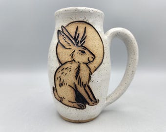 May include: A white ceramic mug with a speckled finish and a single handle. The front features a carved, tan-colored illustration of a rabbit sitting in front of a large, circular moon. The design uses dark outlines to define the rabbit's features.