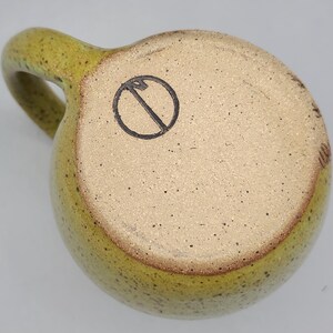 Puede incluir: Una taza de cer&aacute;mica hecha a mano con un esmalte verde oliva moteado. La taza tiene un cuerpo redondeado, un asa resistente y una base de color claro con un s&iacute;mbolo negro. La taza est&aacute; dise&ntilde;ada para bebidas calientes.