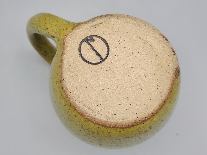 Puede incluir: Una taza de cer&aacute;mica hecha a mano con un esmalte verde oliva moteado. La taza tiene un cuerpo redondeado, un asa resistente y una base de color claro con un s&iacute;mbolo negro. La taza est&aacute; dise&ntilde;ada para bebidas calientes.