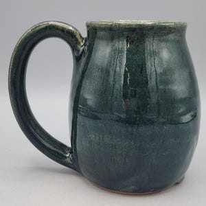 Może przedstawiać: Ceramiczny kubek o zaokrąglonym korpusie i dużym, zakrzywionym uchwycie. Kubek posiada błyszczącą, głęboką leśno-zieloną glazurę z subtelnymi r&oacute;żnicami w odcieniu i fakturze, gładkie wykończenie oraz nieco ciemniejszą krawędź u g&oacute;ry.